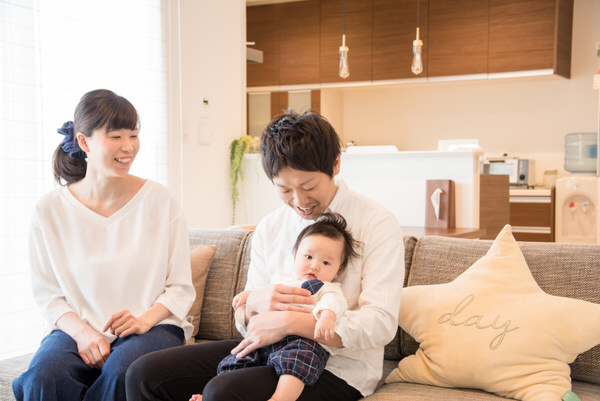 明るいリビングのソファに座り、赤ちゃんを抱っこして微笑む父親と、その隣で見守る笑顔の母親